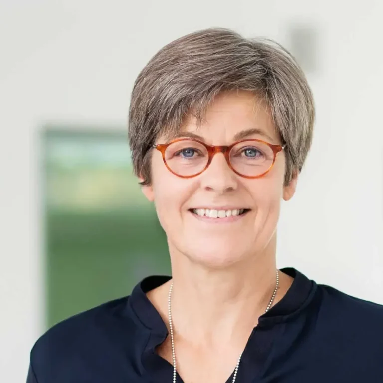 (C) Smart Nanotubes Eine lächelnde Frau mit grauen Haaren und Brille in einem hellen Raum.