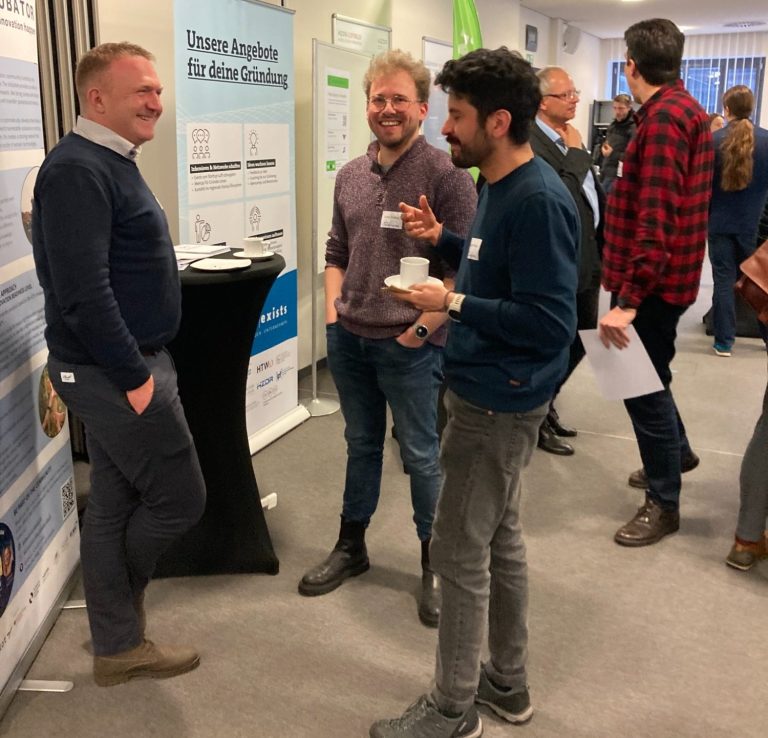 Community (c) SENSCUBATOR Menschen und Stände auf einer Messe, moderne Architektur und Bildschirmpräsentationen.