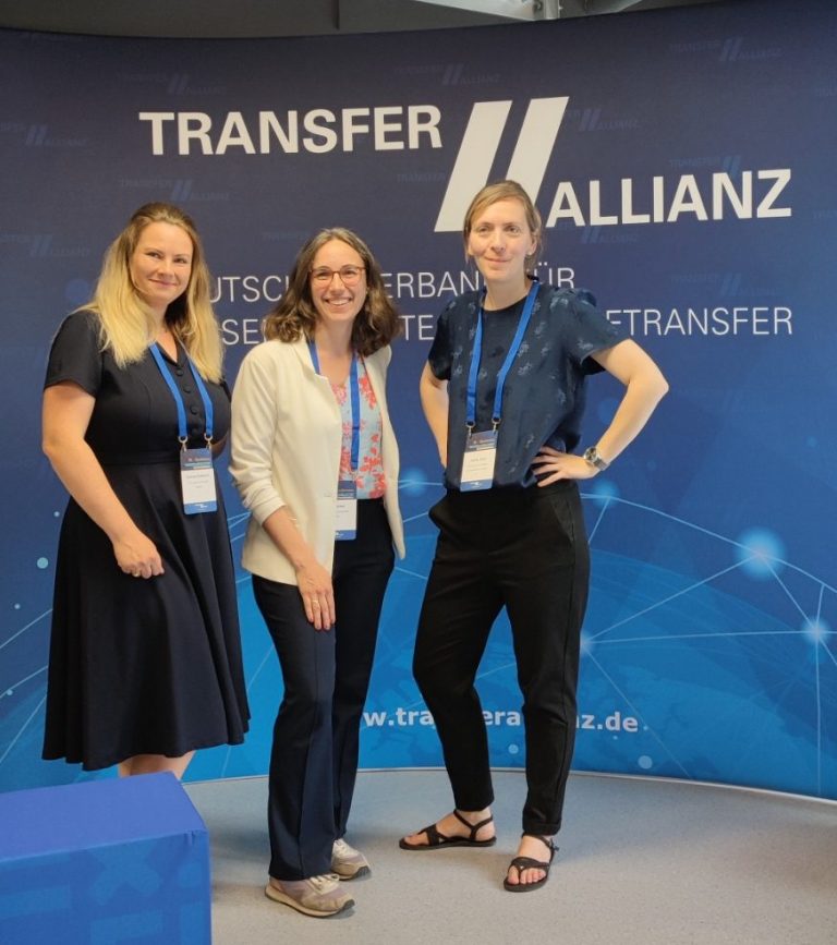 (c) SENSCUBATOR Drei Frauen stehen lächelnd vor einem Banner der Transfer Allianz.