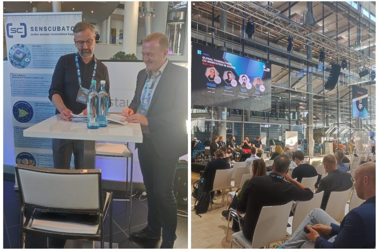 Zwei Männer an einem Stand, im Hintergrund eine Veranstaltung mit Publikum und einer Leinwand.