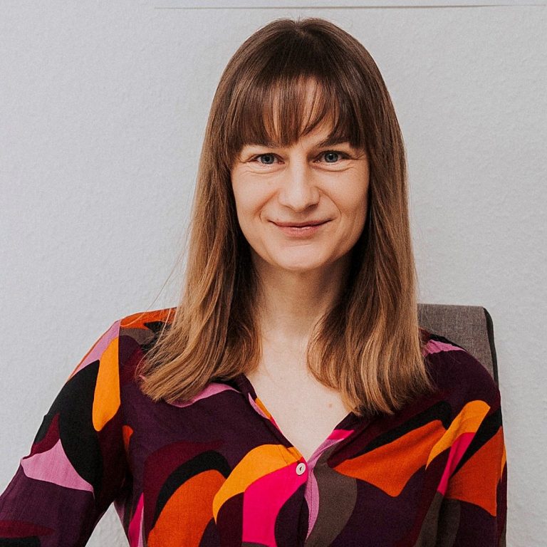 (C) Karin Weigelt Frau mit langen, braunen Haaren in einem bunten, gemusterten Oberteil lächelt.